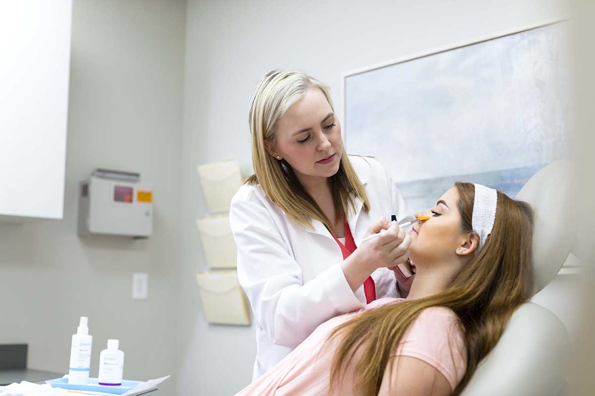 A doctor with her patient laying in the chair eyes closed as the doctor applies solution to her face.