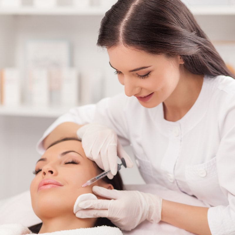 A doctor injecting a solution inside the face of a patient as she lays down the doctor hovers over her with a needle inside her cheek.