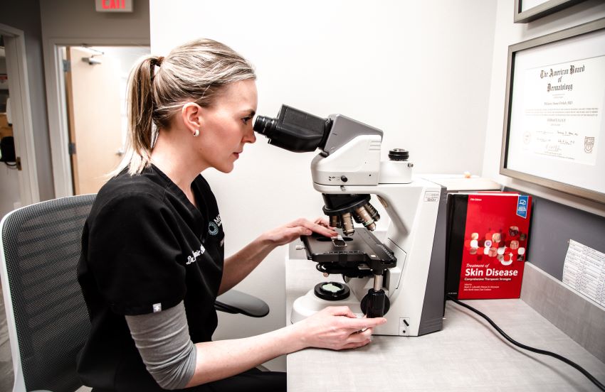female dermatologist sits at desk and looks into microscope